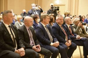 Grupa mężczyzn siedzi na krzesłach w pierwszym rzędzie i patrzy przed siebie.