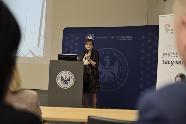 Kobieta stoi za mównicą. W dłoni trzyma mikrofon. W tle duży baner “ Uniwersytet muzyczny w lublinie”.