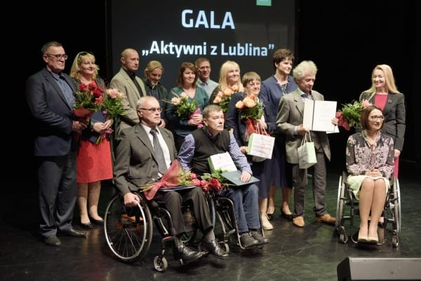 Na scenie pozuje do zdjęcia grupa osób na tle napisu: “ Gala Aktywni z Lublina”. Na przodzie są trzy osoby na wózkach inwalidzkich. Za nimi stoi reszta grupy. Wszyscy stoją lekko odwrócenie w prawą stronę od obiektywu.