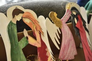 Rękodzieła aniołów. Aniołowie są w różnych kolorowych szatach, jedni mają ręce do góry inni złożone do modlitwy.