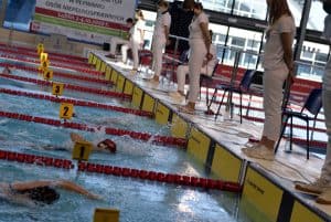 Zawodnicy płyną na swoich torach. Poza basenem stoją kobiety ubrane na biało i obserwują.