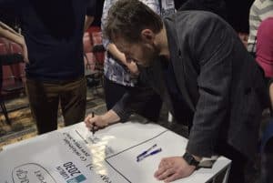 Pochylony mężczyzna w średnim wieku uzupełnia dużą tabelę leżącą na stole. Trzyma marker po prawej stronie tabeli. Drugą ręką opiera się o stół.