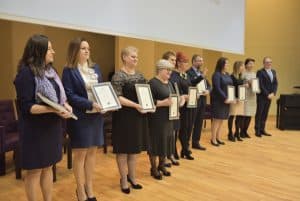 Grupa osób pozuje do zdjęcia na scenie. Wszyscy trzymają przed sobą dyplomy i stoją bokiem do obiektywu.