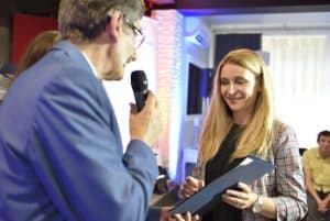 Mężczyzna trzyma w dłoni dyplom i mikrofon. Obok niego stoi kobieta. Jej twarz jest zwrócona w kierunku dyplomu.