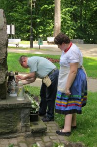 Mężczyzna pochyla się do zniczy. Obok niego stoi kobieta. W tle drzewa parku.