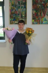 Kobieta w obu dłoniach trzyma kwiaty. Uśmiecha się. W tle kolorowe obrazy na ścianie.