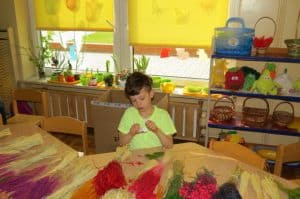 Na stole leżą różnokolorowe gałązki, ususzone kwiaty. Za stołem siedzi mały chłopiec i trzyma klej. Za nim jest parapet i okno, gdzie stoją rośliny.