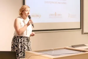 Kobieta trzyma w ręku mikrofon i przemawia. Stoi obok wyświetlanej prezentacji.