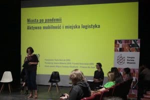Kobieta przemawia do mikrofonu stojąc. Za nią wyświetla się prezentacja pt. “ Miasta po pandemii. Aktywna mobilność i miejska logistyka. Po prawej stronie grupa ludzi siedzi.