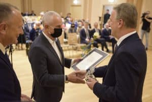 Trzech mężczyzn stoi na scenie. Jeden z nich trzyma oprawiony dokument i wyciąga rękę w geście gratulacji. Drugi mężczyzna trzyma statuetkę i także wyciąga rękę w geście gratulacji. Wszyscy stoją bokiem do obiektywu.