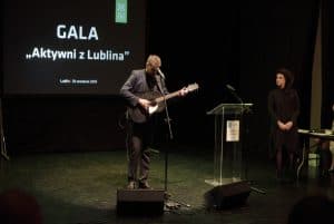 Na scenie jest mężczyzna z gitara elektyrczną, a przed nim jest mikrofon. Po prawej stronie stoi kobieta, która patrzy na mężczyznę. W tle jest wyświetlany napis “Gala Aktywni z Lublina”.