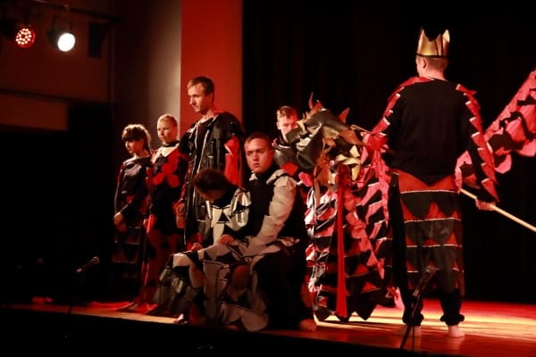 Na scenie jest grupa osób. Wszyscy mają na sobie stroje. Mężczyzna na skraju sceny kuca.