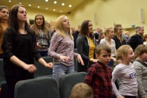 Ludzie stoją na widowni. Ich twarze są zwrócone przed siebie. Niektórzy się uśmiechają.