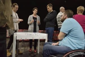 Mężczyzna na wózku jest przez stołem. Jest on sfotografowany bokiem po prawej stronie. Osoby stoją nad białym stołem, na którym znajduje się biała z nagłówkiem Sport.