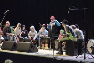 Na scenie jest grupa osób. Pośrodku stoi mężczyzna. Wokół niego siedzą osoby i mają instrumenty.