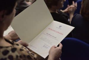 Kobieta w dłoniach trzyma dyplom uznania. Widnieje tam napis: Duplom uznania dla Pani Doroty Anyszek”. Nie jest ona ujęta w pełni w kadrze i jest tyłem do obiektywu.