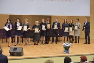 Grupa osób stoi na widowni z dyplomami w rękach. Przed nimi stoi duży bukiet kwiatów. Tyłem do obiektywu siedzi widownia, której widać tylko głowy.