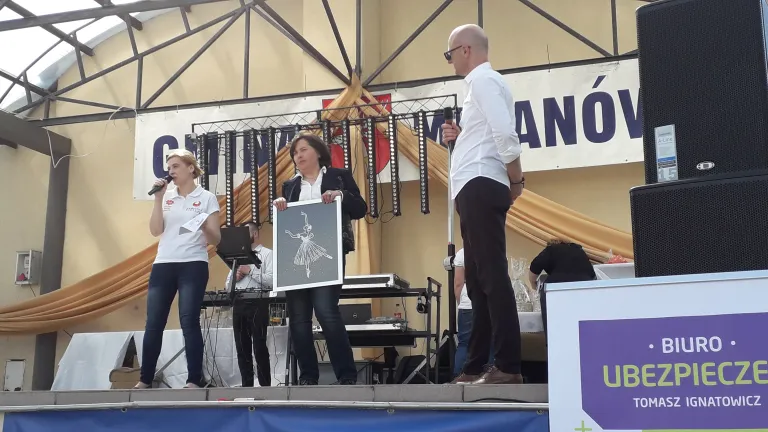 Na scenie stoi grupa osób. Po lewej stronie kobieta ma mikrofon blisko twarzy, a na środku stoi kobieta, która trzyma czarno-biały obraz baletnicy. Po prawej stronie bokiem do obiektywu stoi mężczyzna, który jest odwrócony w kierunku tych dwóch pań.