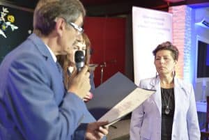 Mężczyzna trzyma w dłoni dyplom i mikrofon. Obok niego stoi kobieta. Jej twarz jest zwrócona w kierunku dyplomu.