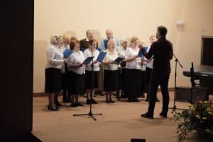 Grupa osób stoi na scenie. Wszyscy trzymają śpiewniki. Przed nimi stoi mężczyzna i ma ręce podniesione.