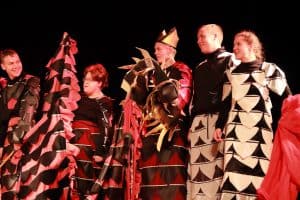 Osoby stoją na scenie. Jeden z mężczyzn ma podniesiona rękę. Wszyscy się uśmiechają.