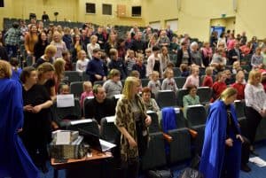 Niektórzy stoją inni siedzą na widowni. Ludzie mają twarze zwrócone ku scenie.