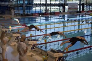 Zawodniczki skaczą do wody. Za nimi stoją osoby ubrane na biało.