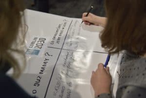 Kobieta na zdjęciu wpisuje słowa w tabelkę po prawej stronie, trzymając długopis w lewej dłoni. Na samej górze kartki widnieje napis Lublin 2030. Pod spodem zaś znajduje się tabelka z nagłówkami, po lewej-” Co mamy?”, a po prawej-” Co chcielibyśmy, żeby było zrobione w 2030?”.