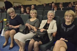 Na widowni siedzą osoby. Kobiety patrzą wprost w obiektyw. KObieta po prawej stronie ma zamknięte oczy.