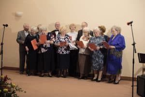 Grupa osób stoi na scenie. Osoby trzymają pomarańczowe śpiewniki.