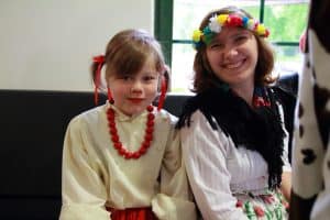 Kobieta i dziewczynka uśmiechają się do obiektywu. Są ubrane w stroje ludowe. Siedzą one na kanapie.