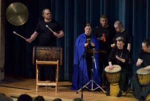 Ludzie stoją na scenie. Niektórzy mają bębenki między nogami. Inny mężczyzna obok siebie ma dzwon i pałeczki do niego.