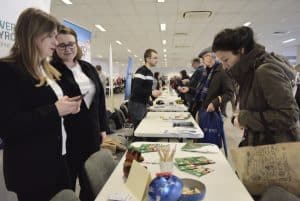 Ludzie pochylają się nad stołami promocyjnymi, na których są ulotki. Jedna z kobiet trzyma w dłoniach smartfona.