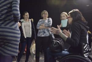 Kobieta na wózku gestykuluje słuchaczkom. Kobiety obok niej mają różne postawy ciała, trzymają założone ręce na piersi lub podpierają swoje podbródki.