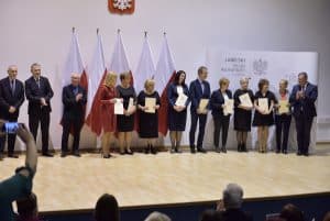 Mężczyźni i kobiety stoją na scenie. Trzymają oni dyplomy w rękach. Mężczyzna po prawej stronie trzyma mikrofon przy twarzy.