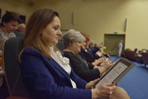 Grupa osób w rzędzie siedzi na widowni. Kobieta trzyma przed sobą dyplom. Za nią starsza kobieta w ręce trzyma okulary.