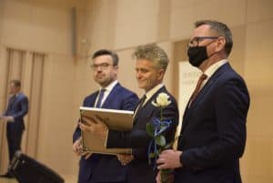 Trzech mężczyzn stoi. Mężczyzna po prawej stronie trzyma białą róże, w środku oprawiony dokument, a z lewej patrzy wprost w obiektyw,