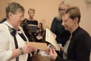 Kobiety stoją naprzeciwko siebie. Po prawej stronie kobieta trzyma kartkę i się uśmiecha. Kobieta po lewej stronie wręcza jej kartkę. Za nimi stoją inne kobiety.