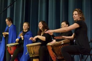Kobiety siedzą na scenie. Między nogami mają bębenki. Uśmiechają się.