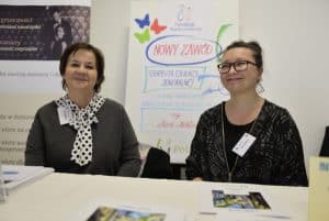 Dwie kobiety siedzą przy stole. Uśmiechają się do zdjęcia. W tle są pionowe banery reklamowe.