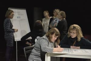 Na pierwszym planie jedna kobieta uzupełnia dużą, białą kartę. Druga kobieta patrzy się na nią. Za nimi grupa osób stoi przy tablicy.