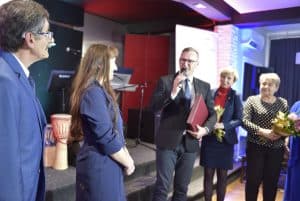 Ludzie stoją na scenie. Jeden z mężczyzn trzyma mikrofon w dłoni i teczkę. Inni trzymają kwiaty.