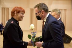 Mężczyzna nachyla się do kobiety. W ręce trzyma białą różę. Kobieta uśmiecha się. Oboje mają złączone ręce w geście gratulacji i stoją bokiem do obiektywu.