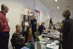 Ludzie stoją i siedzą przy stołach reklamowych. Na stołach są laptopy i ulotki reklamowe. Uśmiechają się do zdjęcia. W tle są pionowe banery reklamowe.