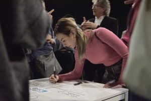 Pochylająca się, młoda kobieta uzupełnia wpis na dużej białej kartce. Kobieta jest ukazana bokiem. Wokół niej stoją inne osoby, niektóre ucięte przez obiektyw.