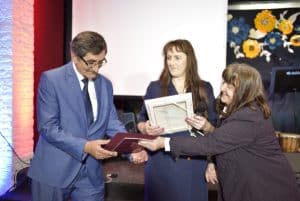 Starsza kobieta wręcza starszemu mężczyźnie teczkę z bordową kartką. W tle za nimi stoi kobieta. Uśmiecha sie.