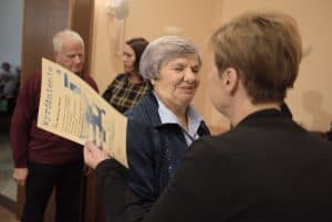 Kobieta stoi tyłem do obiektywu i trzyma kartkę z wyróżnieniem. Przed nią jest starsza kobieta, która się uśmiecha.