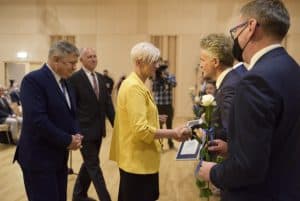 Na scenie jest kilka osób. Kobieta wraz z mężczyna trzymają ręce w geście gratulacji. Za nią jest dwóch mężczyzn. Po prawej stronie stoi mężczyzna i trzyma białą różę.