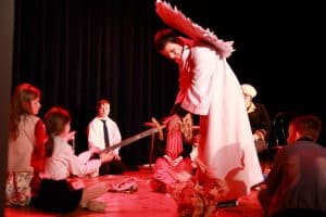 Na scenie jest grupa osób. Wszyscy siedzą w kręgu a w środku jest mężczyzna przebrany za anioła. Trzyma on miecz w dłoni a dziewczynka trzyma dłońmi jego koniec.