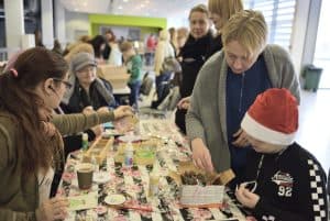 Ludzie pochylają się do stołu z rękodziełami. Chłopiec stojący przy stole ma na głowie czapkę mikołaja.